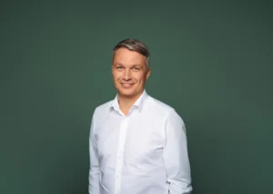 A smiling man with graying hair in a white shirt against a dark green background.