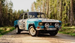 Vintage teal and white rally car parked on a dirt road through a pine forest.