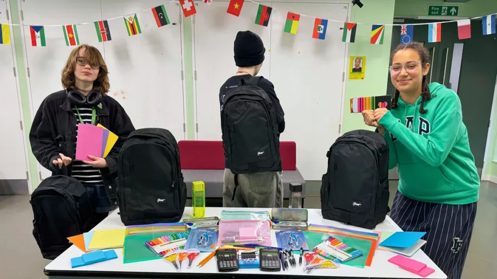 Three students stand behind a table covered with backpacks, stationery, and school supplies.