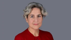 Woman with short wavy grey hair wearing a red top against a solid grey background.