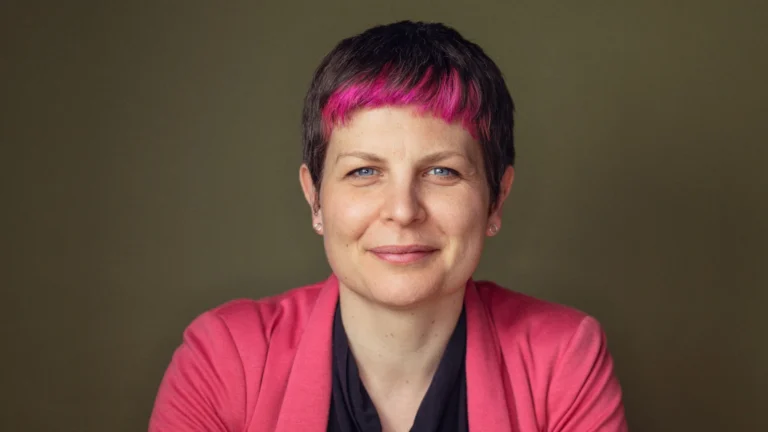 Portrait of a smiling woman with vibrant pink bangs and blue eyes wearing a pink jacket.