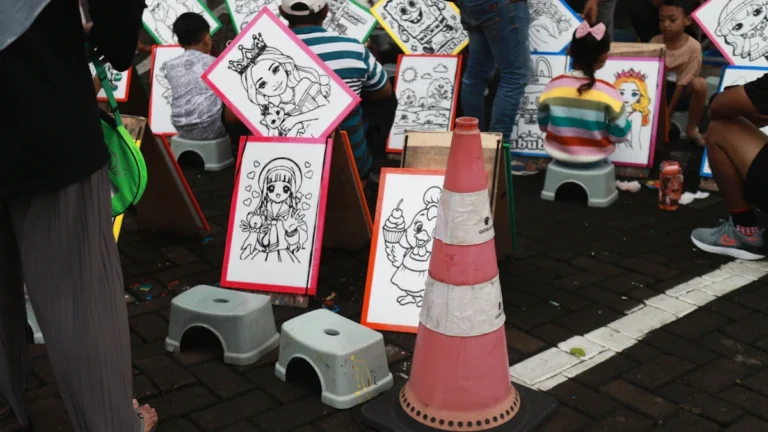 Children and teenagers sitting on stools at an outdoor art station, coloring various cartoon illustrations.