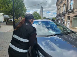 A parking officer in uniform checks a vehicle's windshield on a busy town center street.