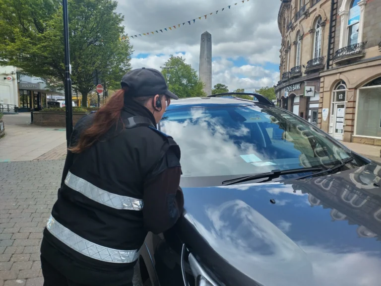 A parking officer in uniform checks a vehicle's windshield on a busy town center street.