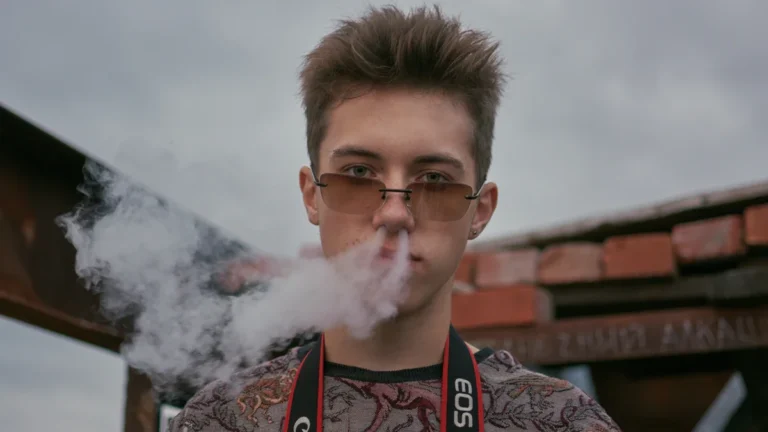 A young man exhaling a cloud of smoke while wearing sunglasses, representing the demographic affected by the new ban.
