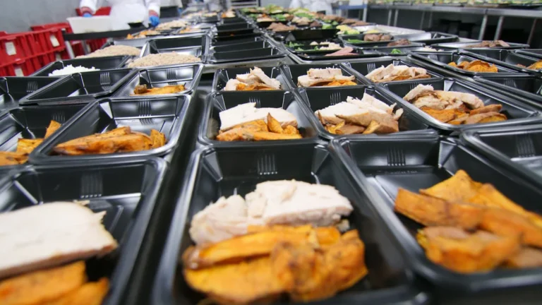 Rows of prepared takeaway meals in black plastic containers on a kitchen assembly line.