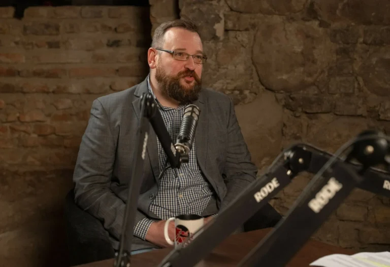 Bearded man in glasses and a grey blazer speaking into a microphone in a studio.