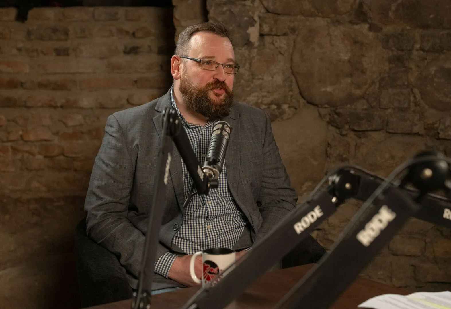 Bearded man in glasses and a grey blazer speaking into a microphone in a studio.