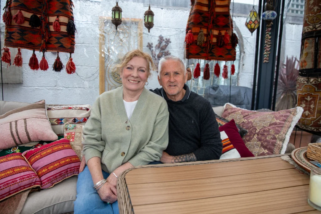 Smiling senior couple sitting together among vibrant woven textiles and colorful patterned cushions.