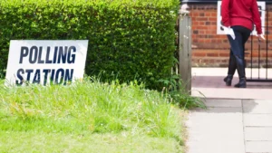 A white sign with bold black text reading POLLING STATION stands before a lush green hedge.