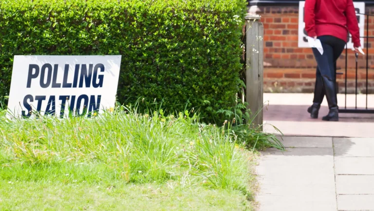 A white sign with bold black text reading POLLING STATION stands before a lush green hedge.