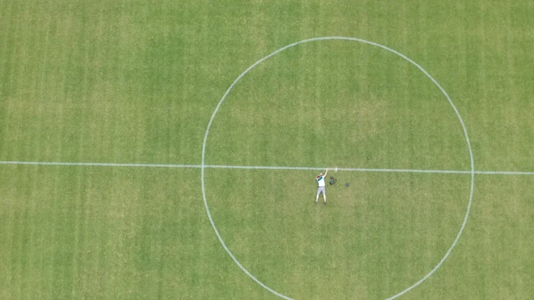 An aerial drone view of a lone man standing in the center circle of a green football pitch.
