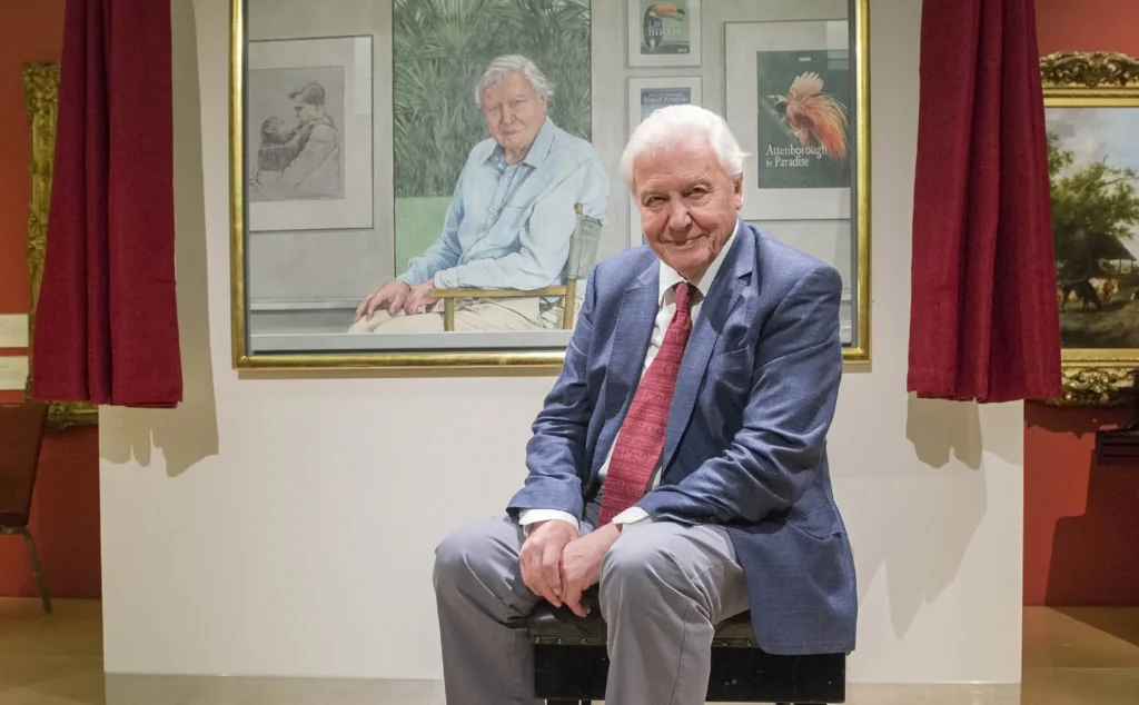 Sir David Attenborough sitting in a blue suit before a large painted portrait of himself.