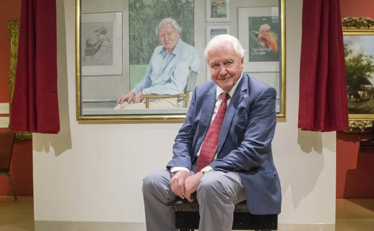 Sir David Attenborough sitting in a blue suit before a large painted portrait of himself.