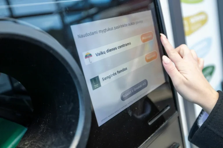 A hand touches a touchscreen on a recycling machine displaying various charity donation options.