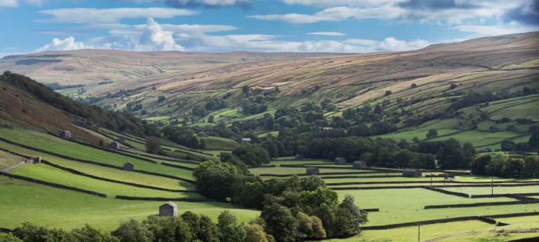 Green rolling hills with stone walls and small stone buildings under a cloudy blue sky.