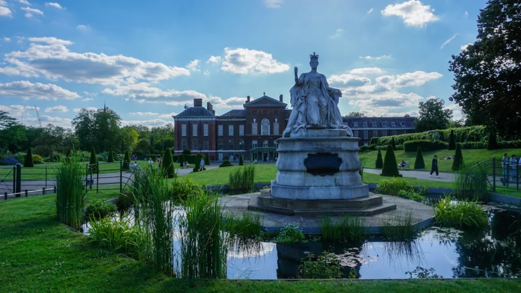 A white marble statue of Queen Victoria seated before her birthplace, Kensington Palace, on a sunny day.