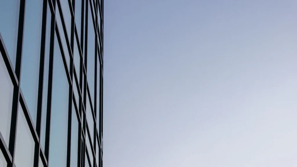 A modern glass building with a clock reflects a clear blue sky on its surface.