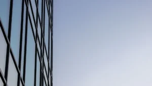 A modern glass building with a clock reflects a clear blue sky on its surface.