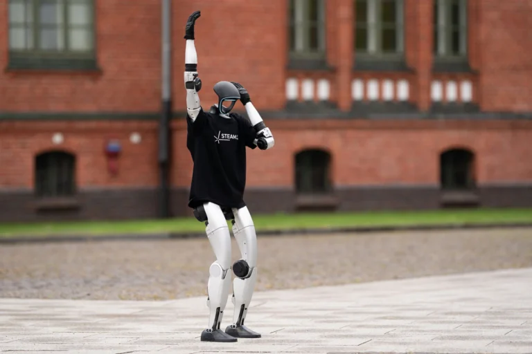 A humanoid robot wearing a black t-shirt raises one arm in front of a brick building.