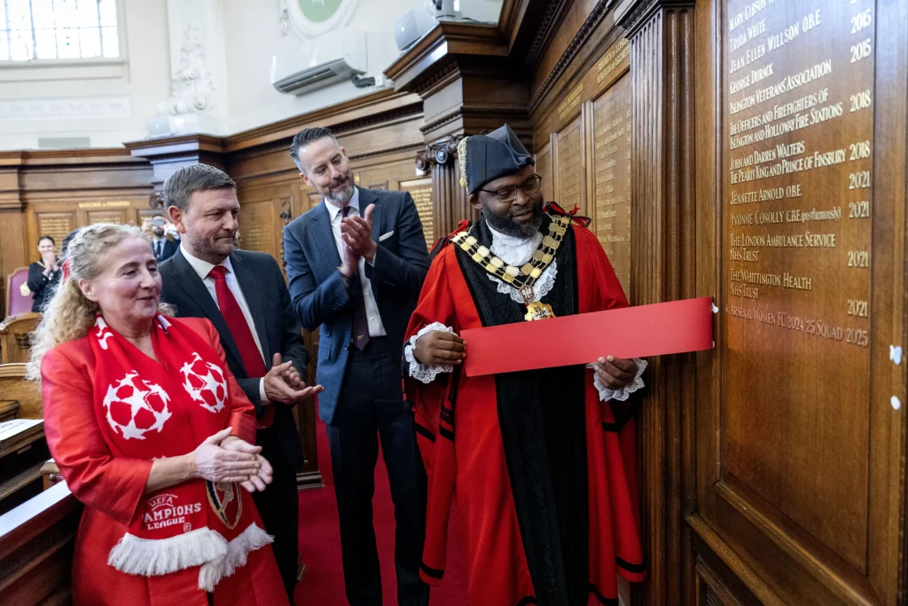 Mayor in ceremonial robes holding a red banner while others applaud near a wooden commemorative wall.