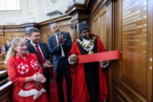 Mayor in ceremonial robes holding a red banner while others applaud near a wooden commemorative wall.