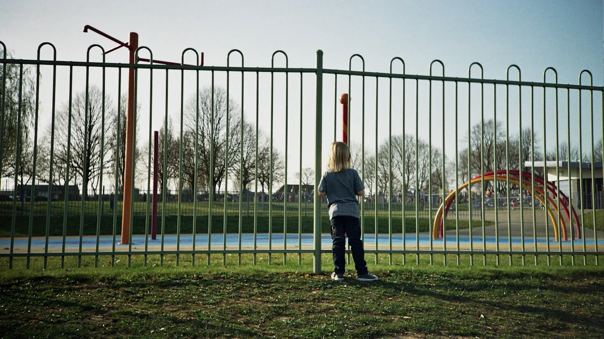 A small child in a blue jacket looking through green metal railings at an empty playground.
