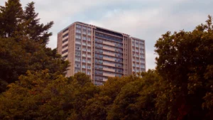 A tall concrete residential apartment block in London rising above a line of green trees.