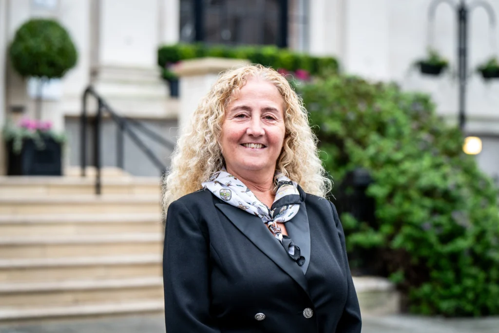 A smiling middle-aged woman with blonde curly hair wearing a black blazer and patterned scarf.