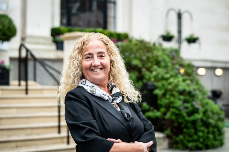 Smiling woman with long curly blonde hair wearing a black blazer and patterned scarf outdoors.