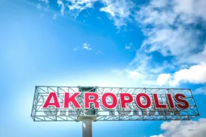 Large red letters spelling 'AKROPOLIS' on a metal framework against a bright blue, cloudy sky.
