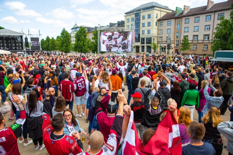Fans in maroon jerseys cheer before a large screen displaying a live hockey match outdoors.