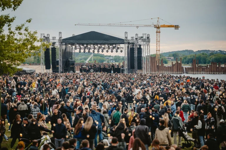 A large crowd of people gathered at an outdoor music stage near a river and crane.