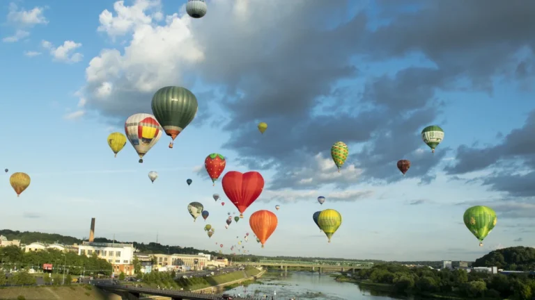 Colorful hot air balloons, including a red heart-shaped one, drift over a river and city skyline.