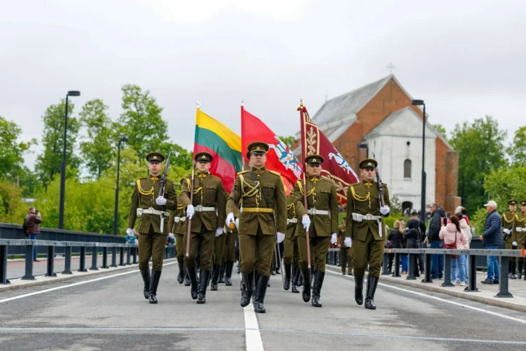 Soldiers in olive-green uniforms march across a bridge while carrying large flags during a ceremony.