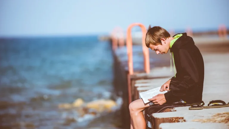 A young person sitting on a concrete pier by the water reading a book in summer.
