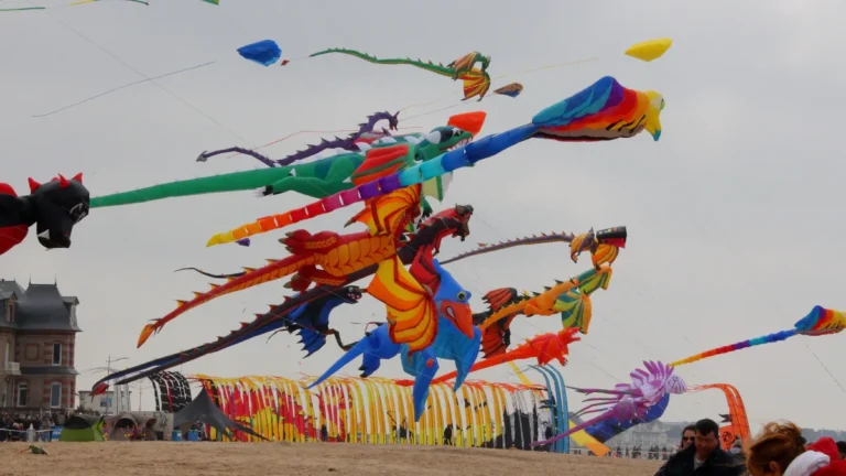 Large, colorful inflatable dragon and creature kites flying over a sandy festival area under an overcast sky.