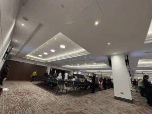 Staff and officials gather at long tables in a large hall for an election vote count.