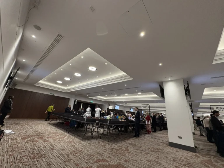 Staff and officials gather at long tables in a large hall for an election vote count.