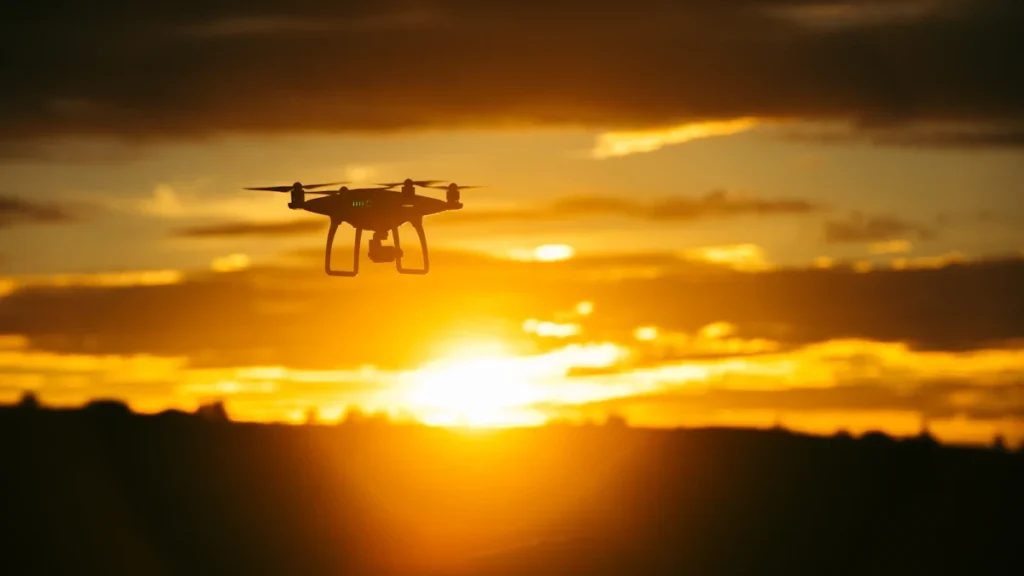 A silhouette of a quadcopter drone flying against a bright orange and yellow sunrise sky.