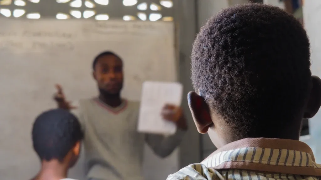 A teacher instructs a class from a whiteboard, captured from the perspective of a student.