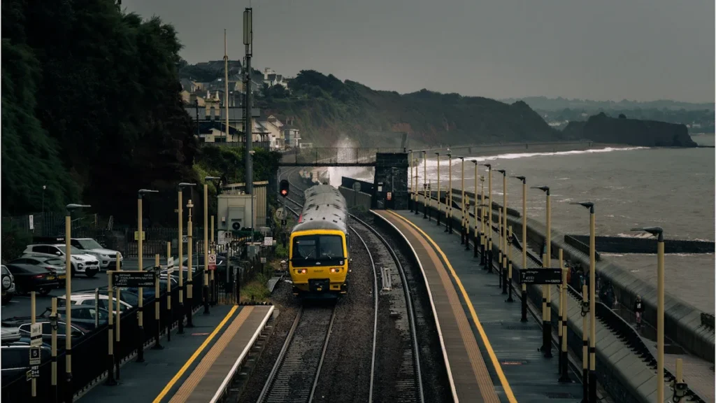 A yellow passenger train approaches a coastal station on a windy day next to the sea.
