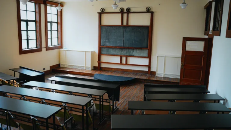 An empty traditional classroom with rows of dark wooden desks and a large blackboard.