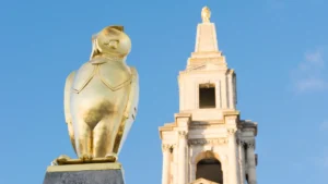 A golden owl statue, the symbol of Leeds, in front of the Leeds Civic Hall tower under a clear blue sky.