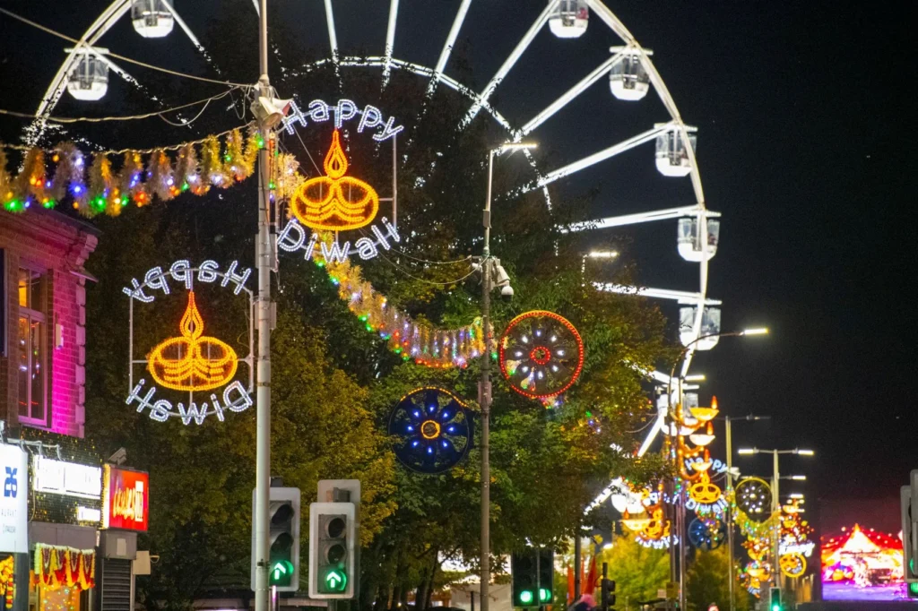 Brightly lit Happy Diwali street decorations stand before a large Ferris wheel at night.