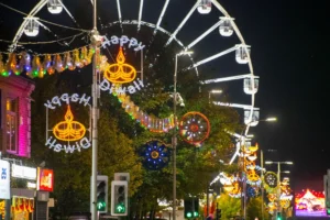 Brightly lit Happy Diwali street decorations stand before a large Ferris wheel at night.