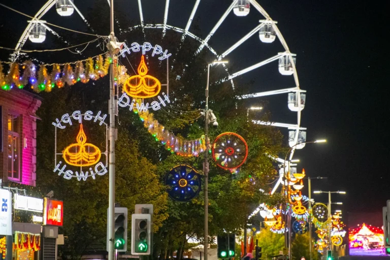 Brightly lit Happy Diwali street decorations stand before a large Ferris wheel at night.