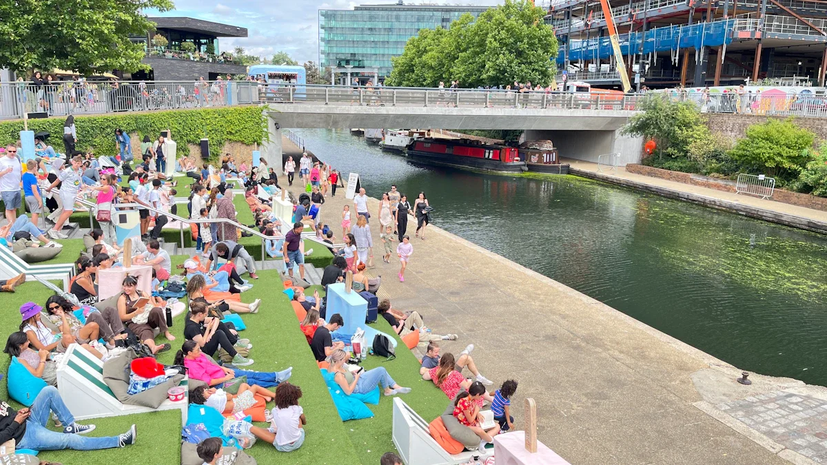 A bustling crowd of people sitting on a grassy bank and walking along a city canal during a summer festival.