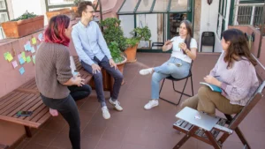 A group of diverse residents sitting in a circle on an outdoor terrace during a collaborative wellbeing support session with sticky notes on the wall.