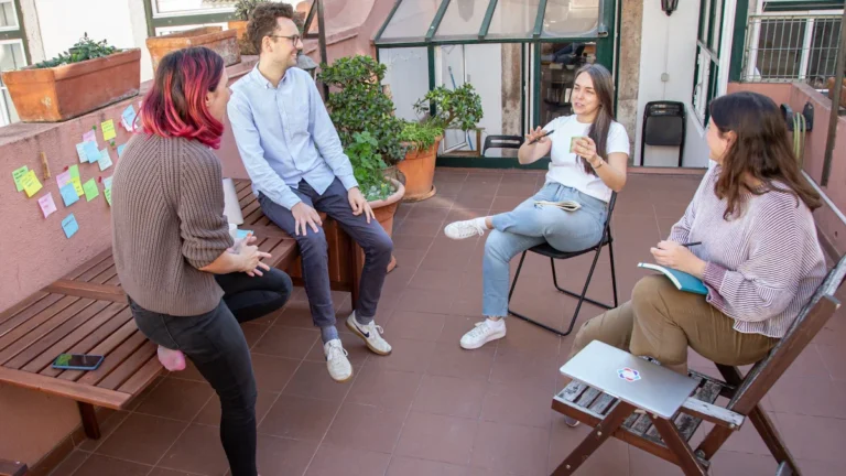 A group of diverse residents sitting in a circle on an outdoor terrace during a collaborative wellbeing support session with sticky notes on the wall.